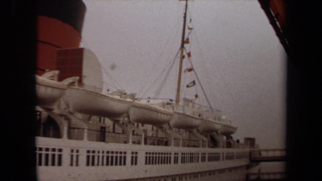 Side of a docked passenger ship with lifeboats, rigging, flags, and a red-and-black smokestack.