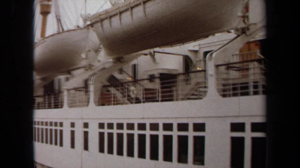 1971 Long Beach home-movie frame of the ship's white side, showing a lifeboat above railings, deck openings, and rows of dark windows.