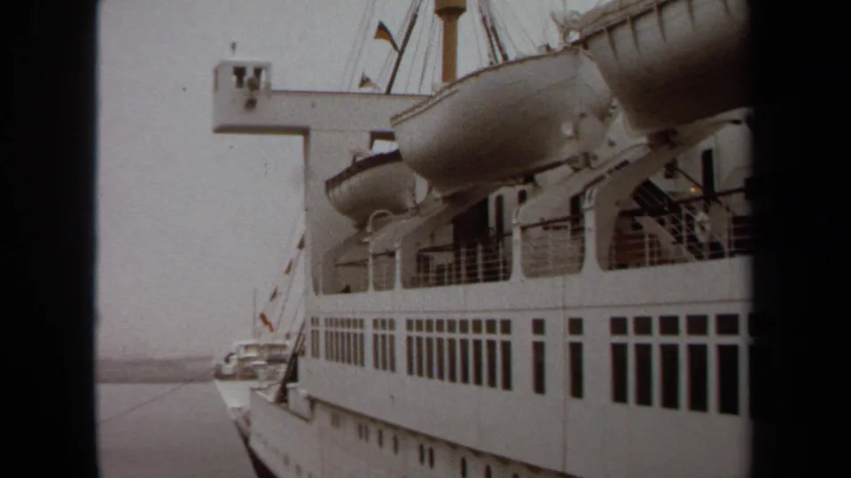 1971 Long Beach frame showing the side of a large white passenger ship with lifeboats, railings, rectangular windows, and a partial harbor setting.