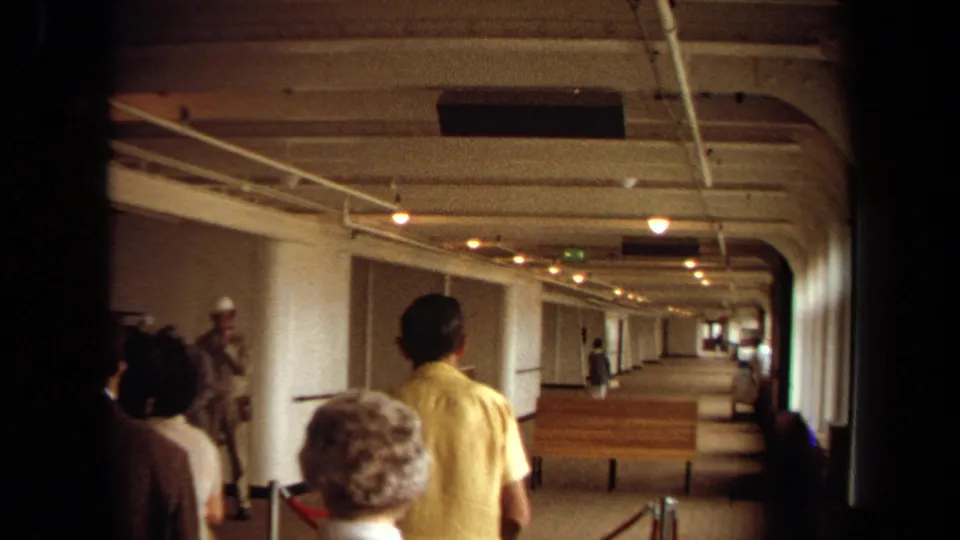 Travelers in 1970s attire move through a Long Beach cruise boarding corridor with benches, stanchions, and warm ceiling lights.