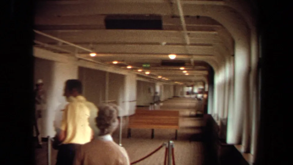 A man and an older traveler walk through the Long Beach cruise boarding corridor, with benches, windows, and stanchions visible.
