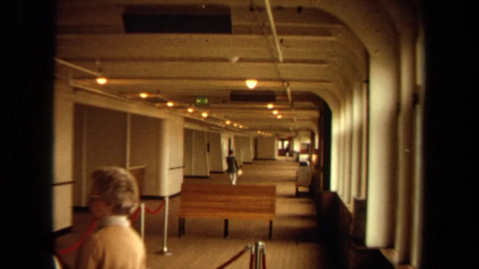 Warm 8mm view down a Long Beach cruise boarding corridor with benches, rope stanchions, windows, and a distant traveler.