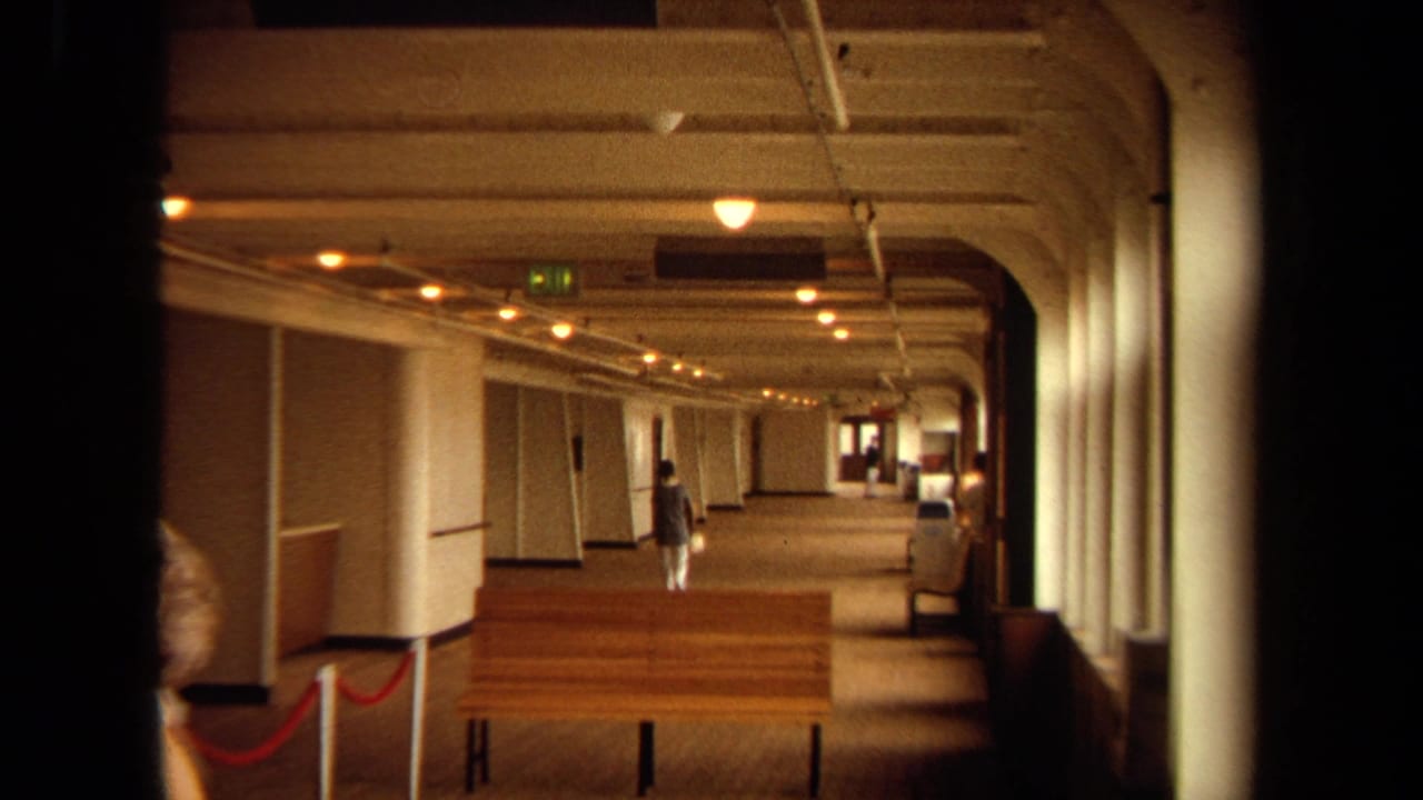 Long carpeted corridor with arched ceiling bays, warm lights, benches, and a woman walking away.
