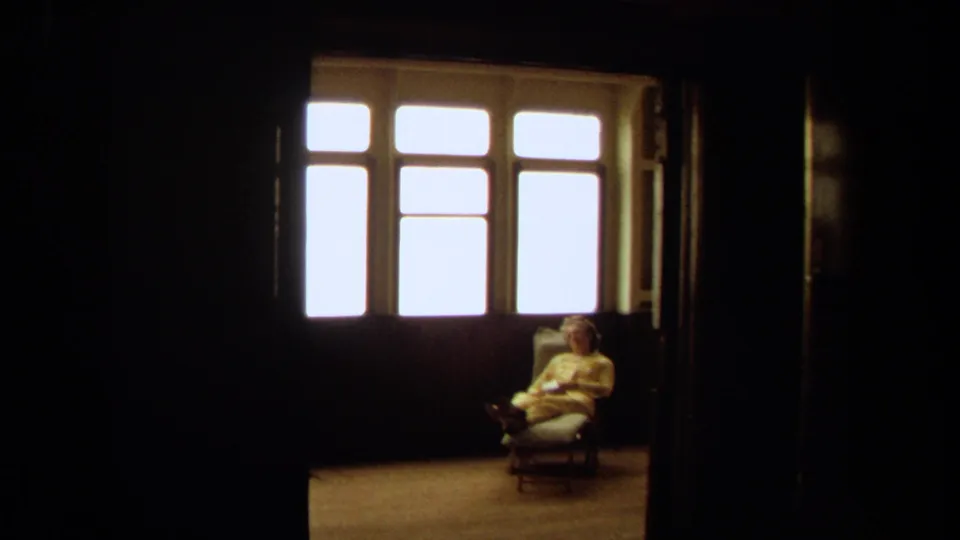 1971 Long Beach interior frame showing an elderly woman seated by a bright window, partially distant in a dark room.