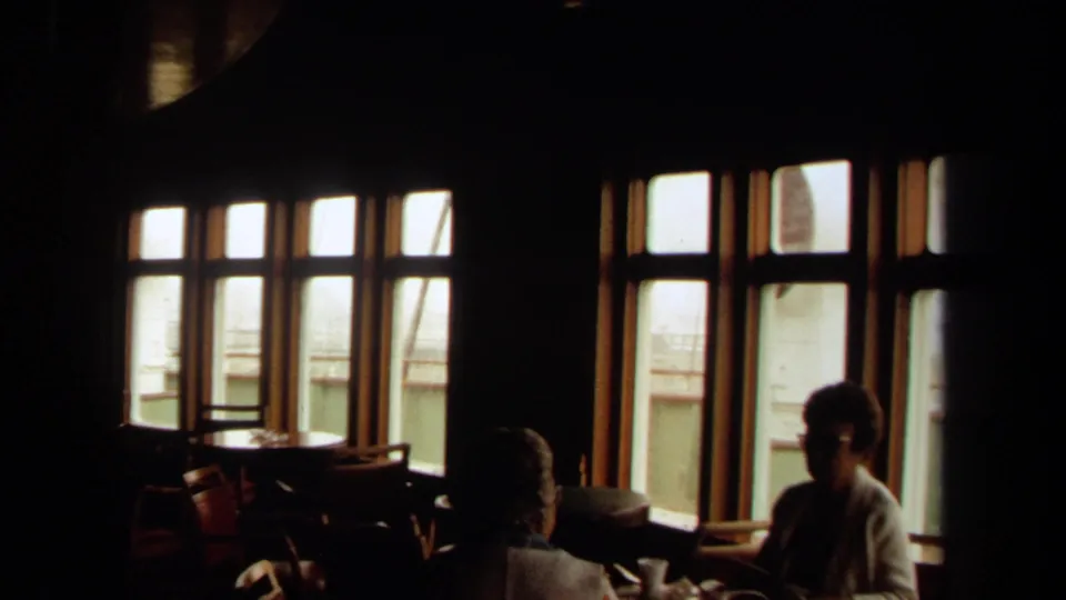 1971 Long Beach home-movie frame showing a dark ship interior, seated adults, tables, chairs, and bright windows looking toward the deck.