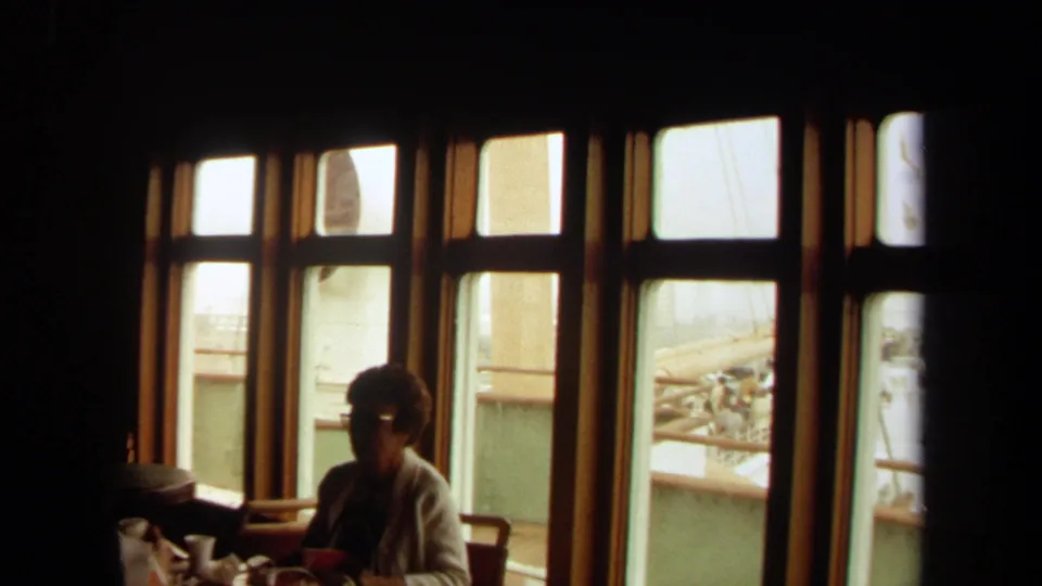 1971 Long Beach 8mm frame of a woman seated in a shadowed ship interior beside large windows overlooking railings and exterior structure.