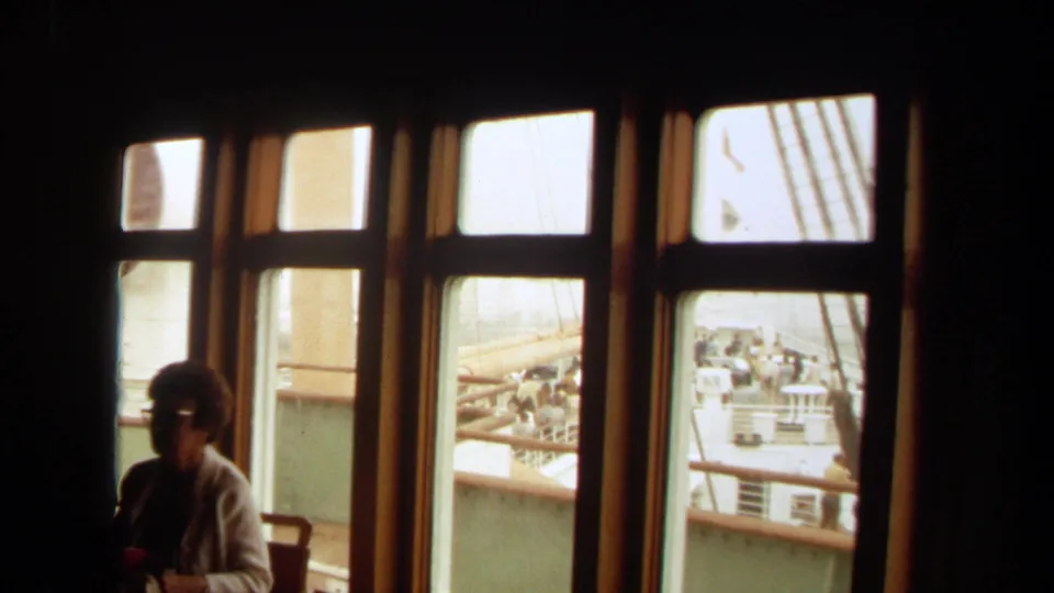 1971 Long Beach home-movie frame showing a seated passenger in a dark ship interior, with windows opening onto railings, rigging, and people outside.