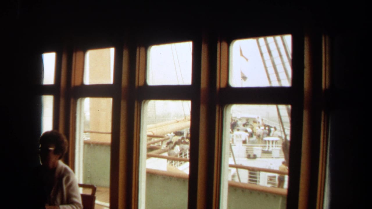 Wood-framed ship windows with deck railings outside and a seated woman in shadow at left.