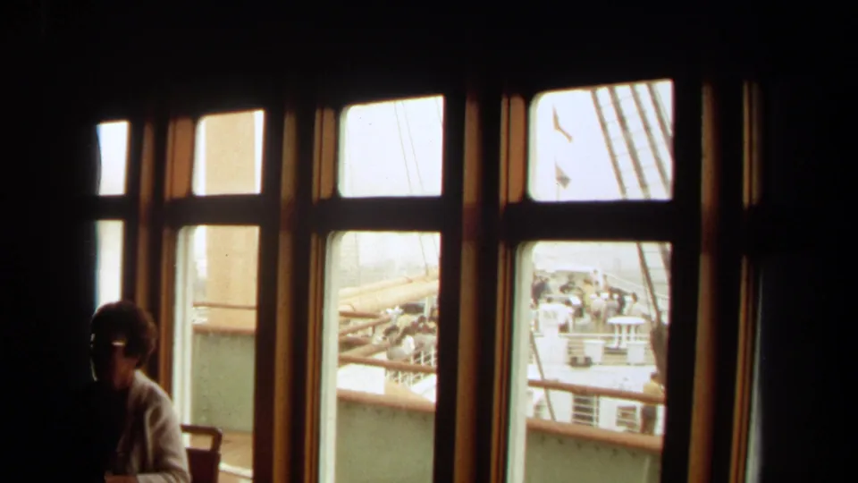 1971 8mm cruise-ship interior view from Long Beach metadata, showing windows, deck railings, distant passengers, and a woman in silhouette.