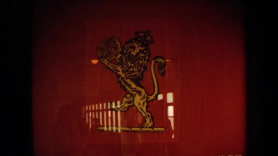 Close view of a red Cunard House Flag display aboard the cruise ship, with a gold lion holding a globe and window reflections across the panel.