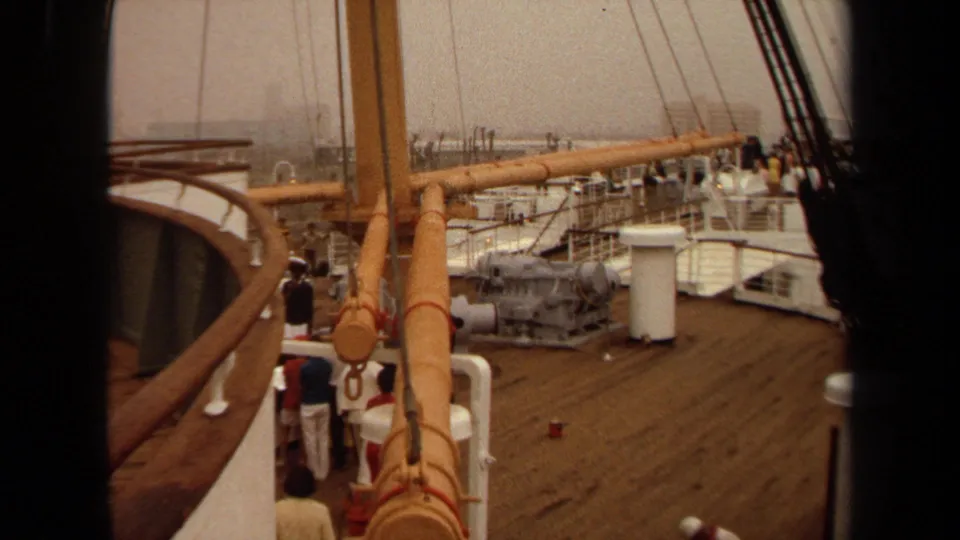 1971 home-movie view of passengers on a ship deck, with wooden railings, rigging, equipment, and hazy harbor structures beyond.