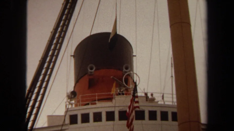 1971 Long Beach home-movie frame of the ship's funnel, deck railings, rigging, mast, and U.S. flag against a pale sky.