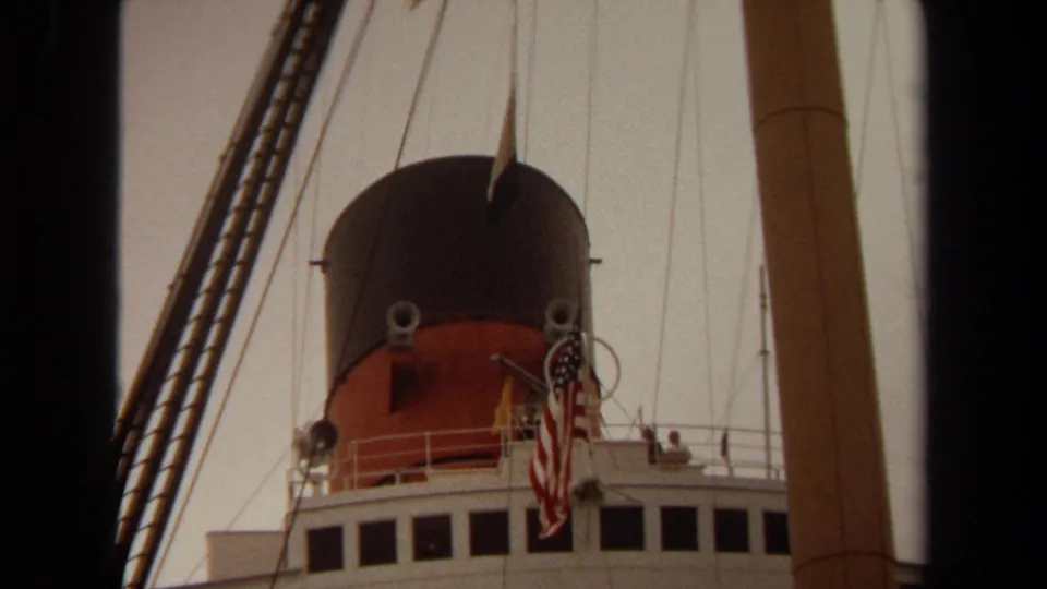 1971 Long Beach home-movie frame looking up at a ship funnel, U.S. flag, rigging, bridge windows, and railings.