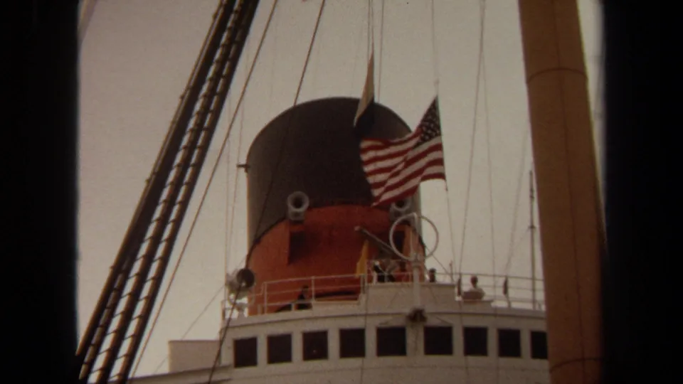 1971 Long Beach 8mm frame showing the U.S. flag fluttering near a ship funnel and upper deck railings.