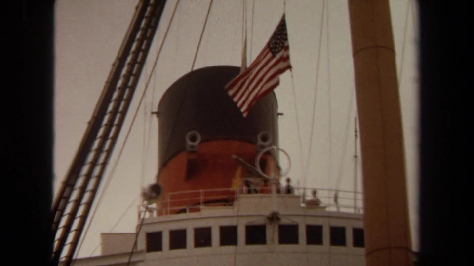 1971 Long Beach home-movie view of a large ship's funnel and U.S. flag through rigging and a vertical mast.