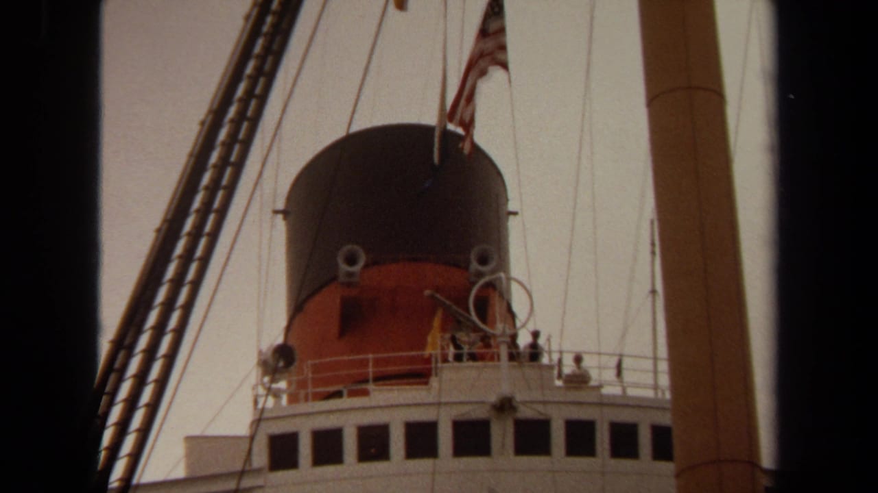 1971 8mm view of the Queen Mary in Long Beach, showing the red and black funnel, American flag, rigging, and deck structures.