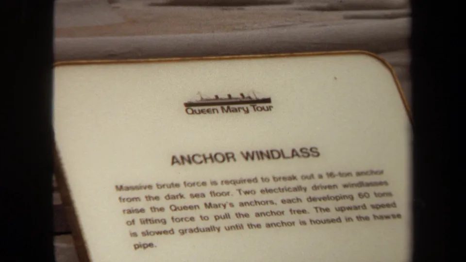 Queen Mary Tour sign describing the anchor windlass, with several visitors visible in the background on the ship deck.