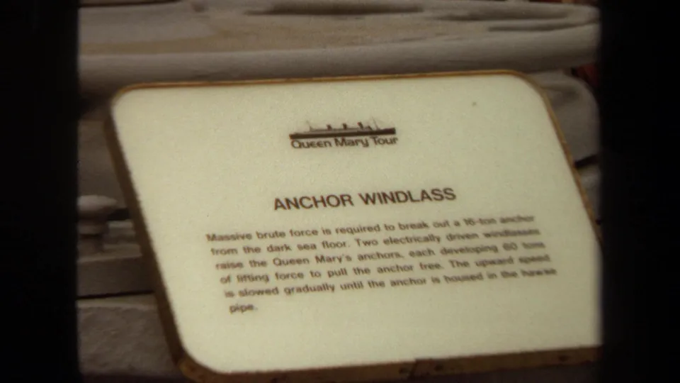 Close 8mm view of the Queen Mary anchor windlass exhibit sign, with deck machinery and ship surfaces around it.