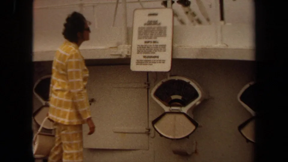 Woman in period yellow checkered clothing stands beside shipboard gauges and signs in Long Beach footage.