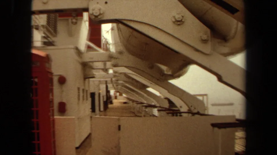 1971 8mm shipboard detail in Long Beach showing stairs, a large support arm, railings, and a red phone booth-like structure.