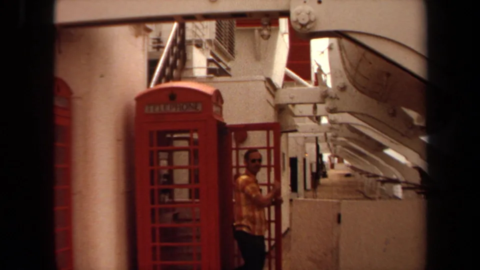 On the 1971 cruise ship deck, a man in a yellow plaid shirt and sunglasses emerges beside the red telephone booth.