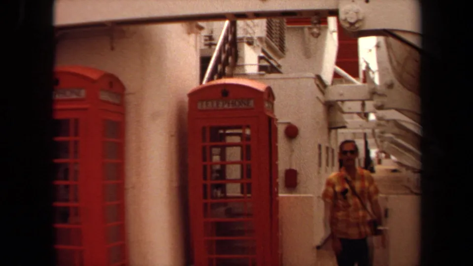 The man walks along the 1971 cruise ship deck past red telephone booths, white railings, and heavy structural beams.
