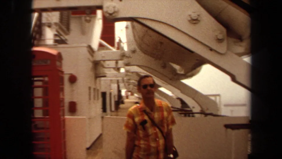 1971 shipboard frame from Long Beach showing a plaid-shirted man near red telephone booths and deck equipment.