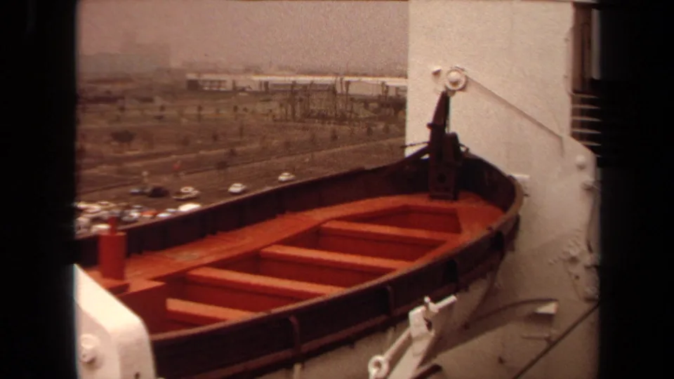 1971 Long Beach shipboard view of a wooden lifeboat mounted beside the deck, with harbor traffic and buildings in the distance.