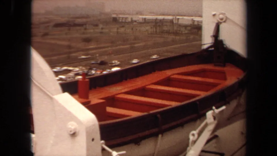 1971 Long Beach home-movie frame showing a mounted ship lifeboat, white davit hardware, harbor roads, and distant buildings.