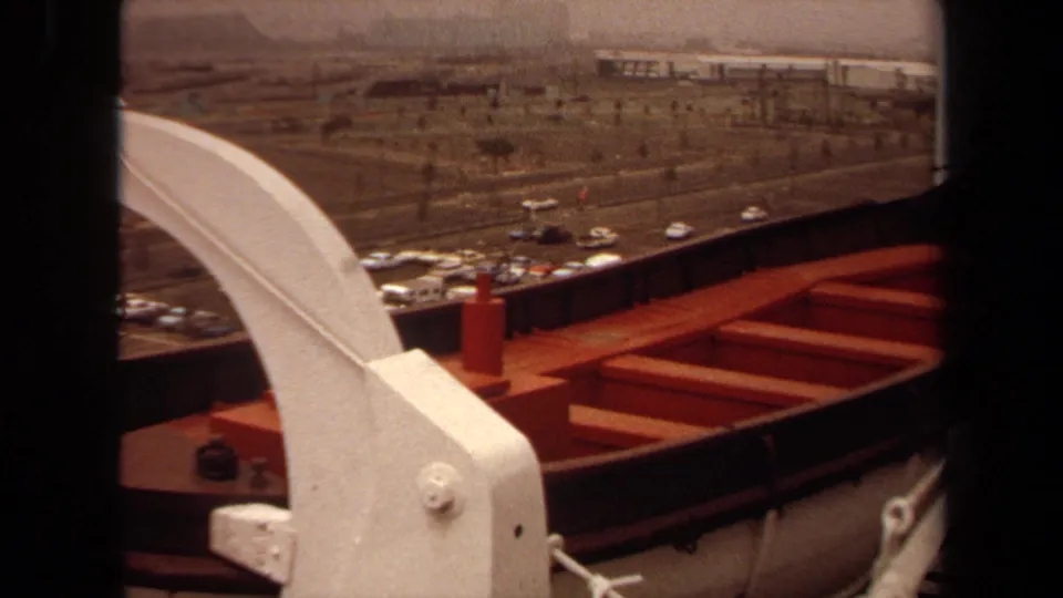 1971 Long Beach ship deck view with a white davit, red lifeboat, rail, roadway, parked cars, and distant port structures.