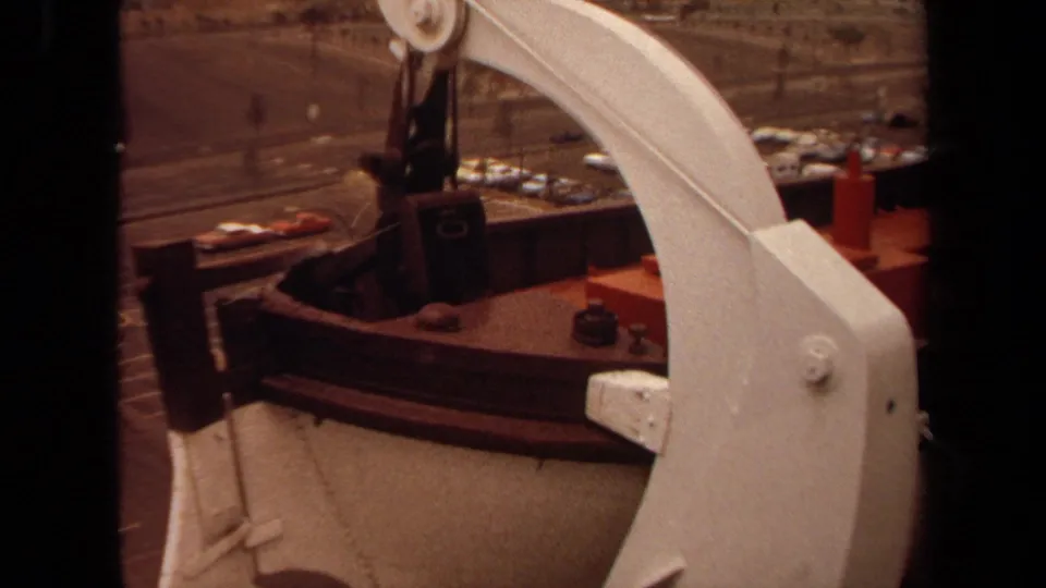 1971 Long Beach shipboard frame with the white davit dominating the foreground and a red lifeboat, rail, cars, and dockside road behind.