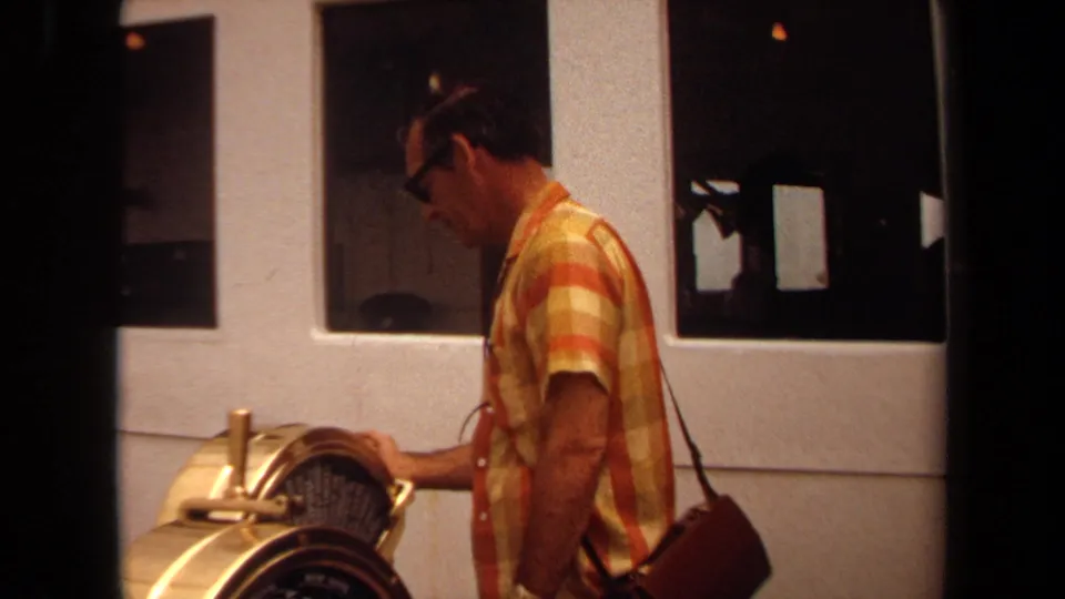 1971 Long Beach frame of an adult man in glasses and a yellow-orange plaid shirt operating a brass-toned industrial machine.