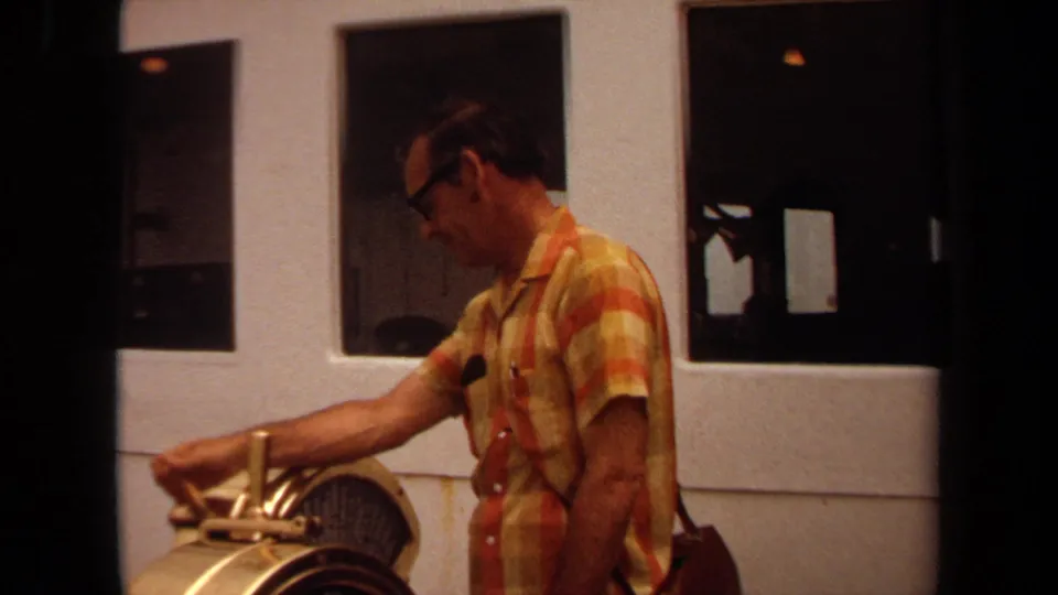 1971 Long Beach 8mm frame showing a man reaching across a brass-toned machine, with dark interior windows behind him.