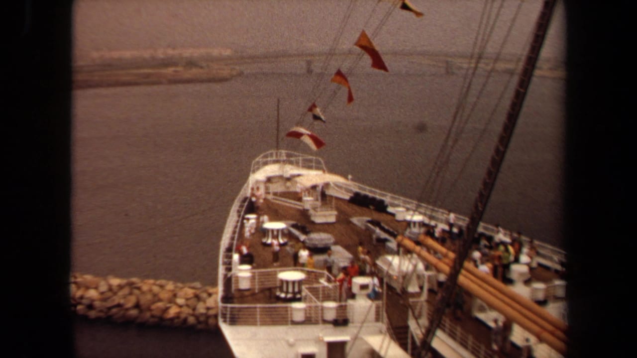 View down onto a ship deck with railings, tables, flags, passengers, water, and breakwater.