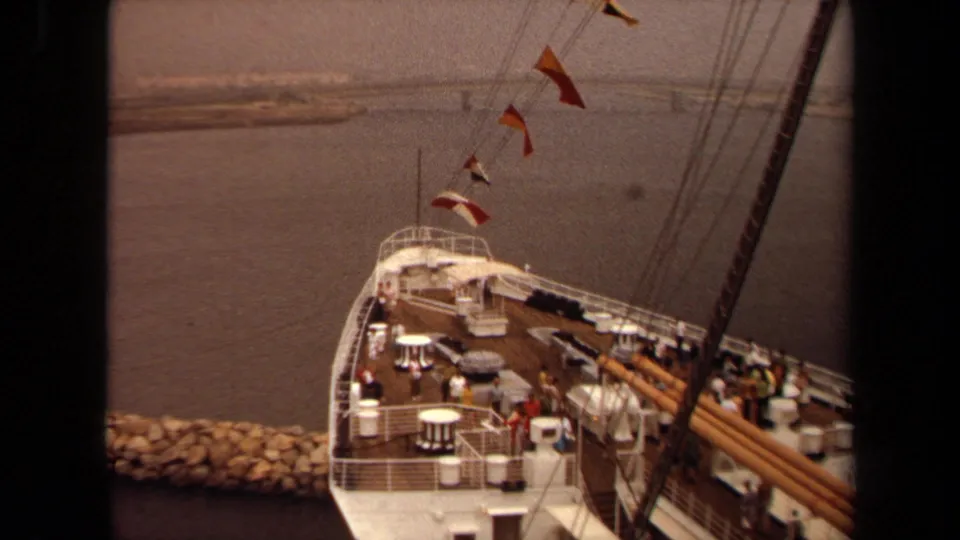 High-angle 1971 Long Beach view of a vessel deck with white railings, small tables, rigging, pennant flags, passengers, harbor water, and breakwater.