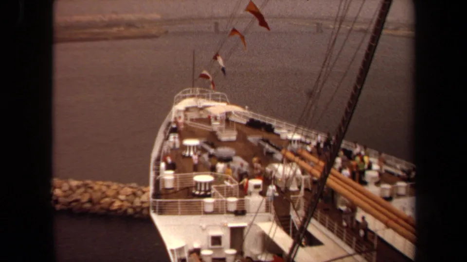Angled 1971 Long Beach deck view showing rigging, pennant flags, white railings, passengers, harbor water, and the rock breakwater beyond.
