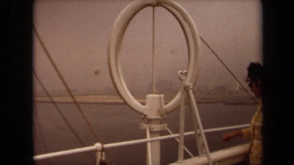1971 Long Beach home-movie frame with a man near the edge of the shot, white ring antenna, deck railing, and hazy water.