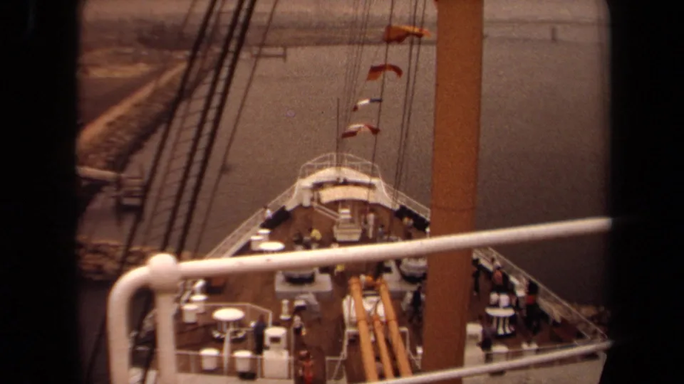 Elevated 1971 Long Beach home-movie view of a passenger ship bow, showing deck fittings, railings, rigging, flags, and distant shore.