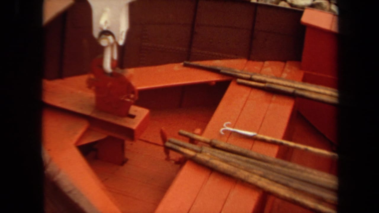 1971 Long Beach 8mm frame showing a red boat deck, wood planking, metal rods, and white lifting hardware near a rocky shoreline.