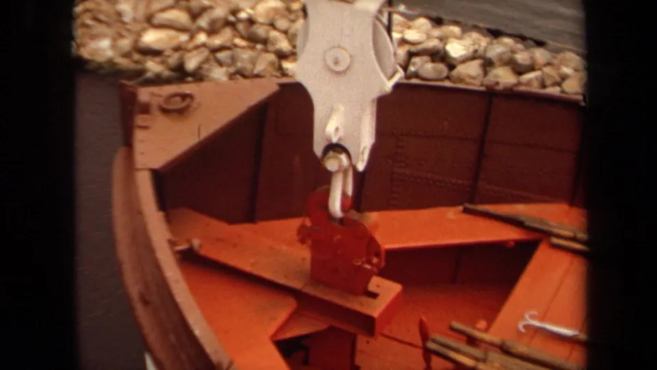 1971 Long Beach harbor frame with a red-painted boat section, white hook assembly, wood panels, metal rods, and shoreline rocks.