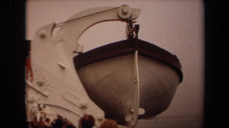 1971 Long Beach frame of a suspended lifeboat beside white ship structure, with davit hardware overhead and bystanders at the bottom.