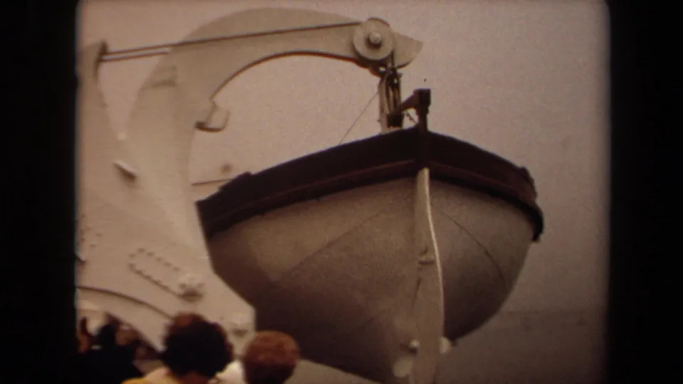 Close 1971 Long Beach 8mm view of a lifeboat descending from a ship davit, with partial spectator heads along the lower frame.