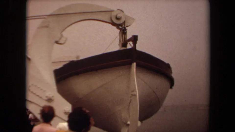 1971 Long Beach home-movie still of a lifeboat lowered beside a ship, framed by davit cables and partial figures at the bottom.