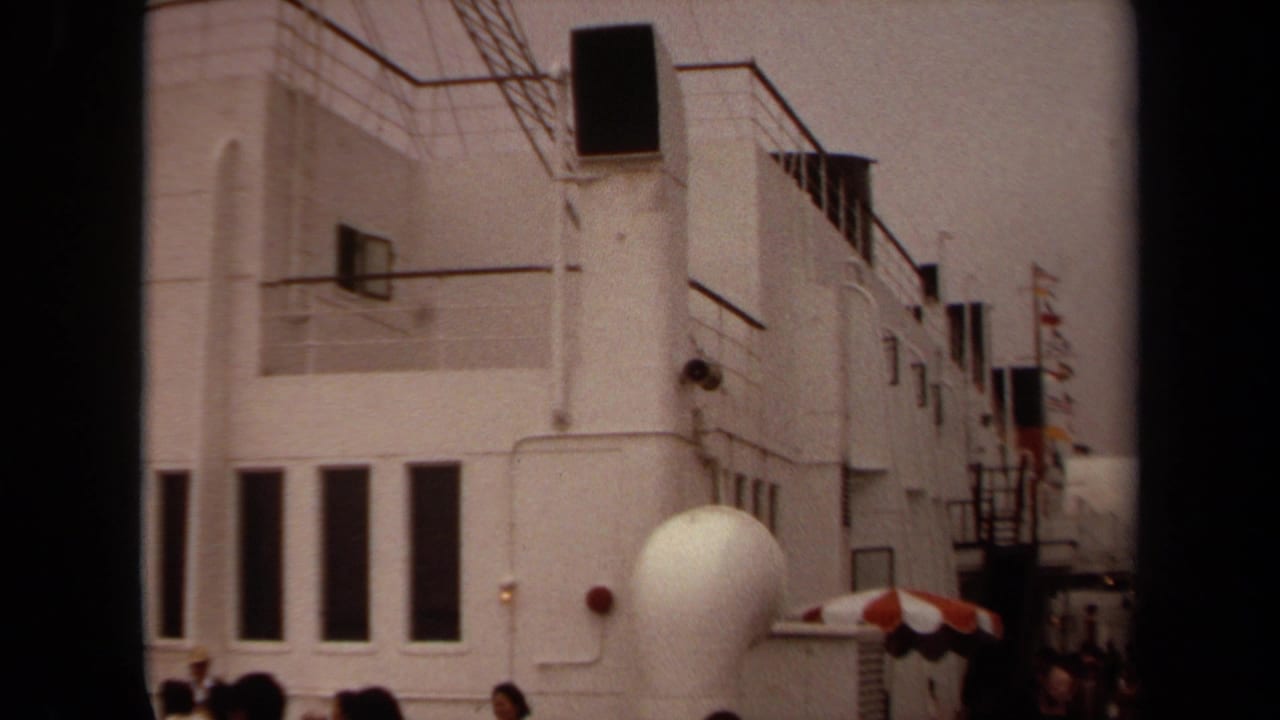1971 Long Beach home-movie frame showing a white Art Deco-style exterior with railings, dark vertical windows, visitors, and a red-and-white umbrella.