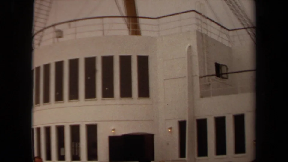 1971 Long Beach home-movie view of a white curved facade with repeated vertical windows, a dark entrance, railings, and visitors at the bottom.