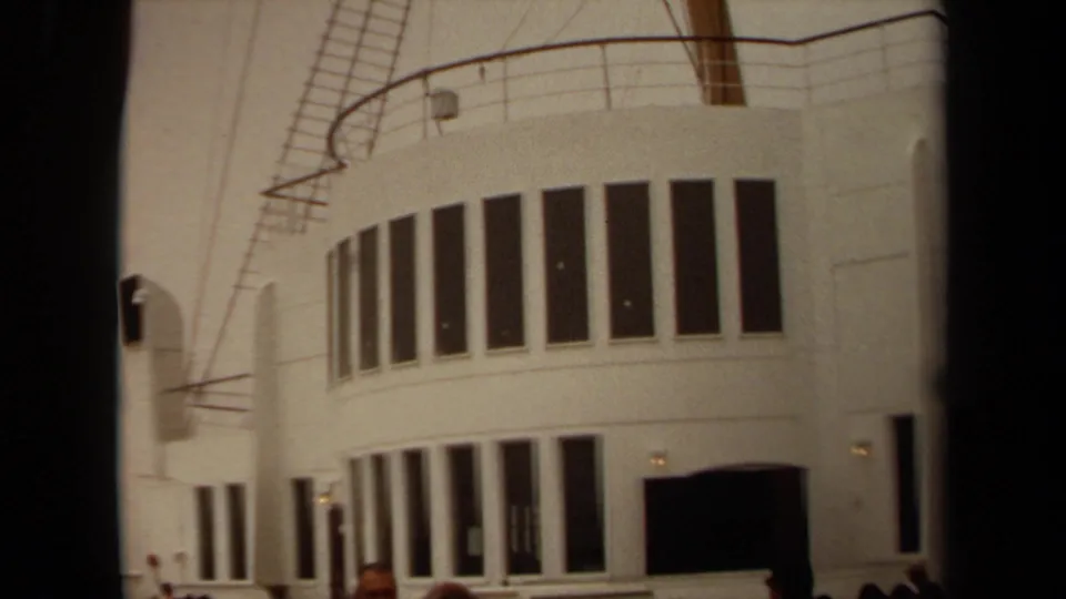 1971 Long Beach frame showing the white rounded building front, repeated dark windows, rooftop railings, small visitors, and warm 8mm color.