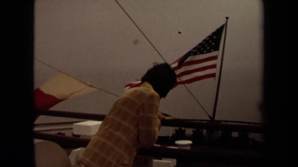 1971 Long Beach shipboard frame with the woman near deck railings and an American flag flying behind her.