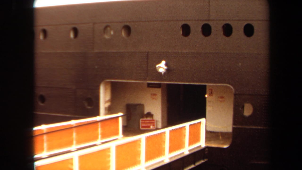 1971 Long Beach 8mm frame showing a docked ship's riveted hull, portholes, orange gangway barriers, and open entry area.
