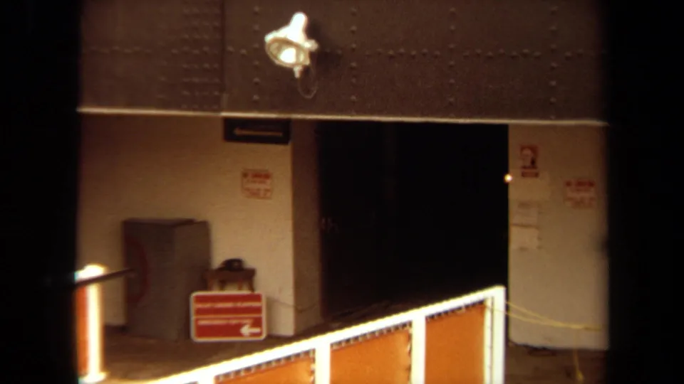 1971 Long Beach 8mm frame of a docked ship's entry area, with orange gangway barriers, red signs, and a dark interior doorway.