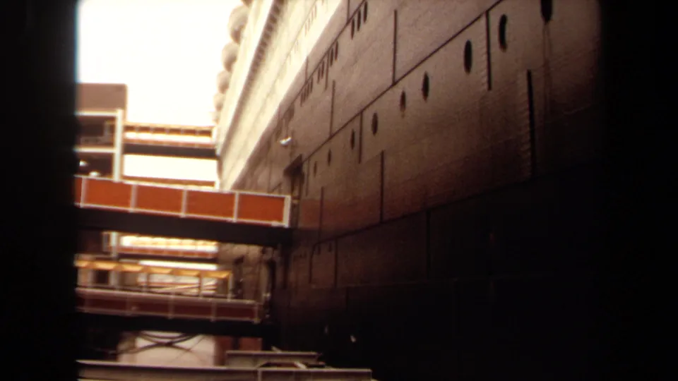 1971 Long Beach view along the dark side of a docked ship, showing portholes, riveted panels, and an elevated orange-railed walkway.